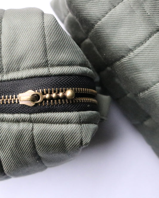 Close-up of two quilted olive green fabric pouches, highlighting their textured stitching and brass zipper with a decorative pull. The fabric has a subtle diagonal weave pattern, and the pouches rest on a white surface.