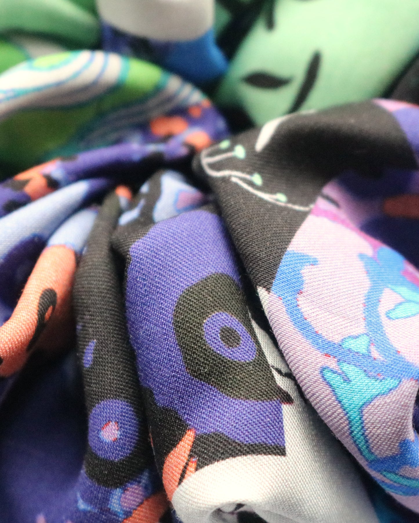 An extreme close-up of a scrunchie made from deadstock fabric, highlighting its vibrant multicolored pattern. The design includes shades of blue, black, purple, green, and orange with abstract and floral elements. The intricate folds and texture of the fabric are visible, showcasing the materialās detail and softness. The background is blurred, emphasizing the fabricās patterns and colors.