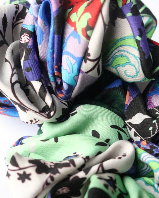 A close-up view of a scrunchie made from deadstock fabric, showcasing its intricate multicolored pattern. The fabric features shades of green, blue, red, black, and white with abstract and floral elements. The ruffled texture and folds of the scrunchie are highlighted, emphasizing the softness and detail of the material. The background is a neutral white surface.